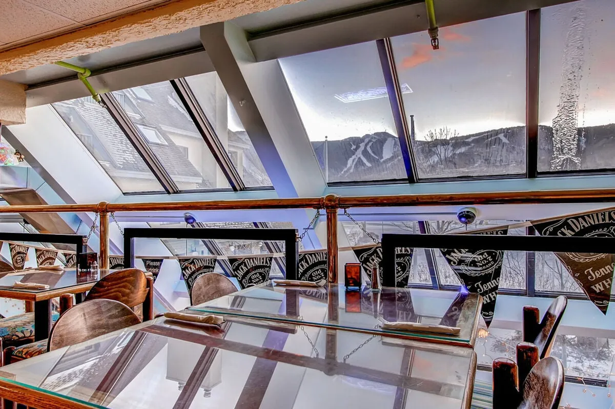 This dining area boasts large windows with scenic mountain views, wooden tables and chairs, and cheerful decorative touches. The open, sunlit setting bridges the kitchen to the communal dining experience, illustrating the ease of serving and sharing meals with loved ones.