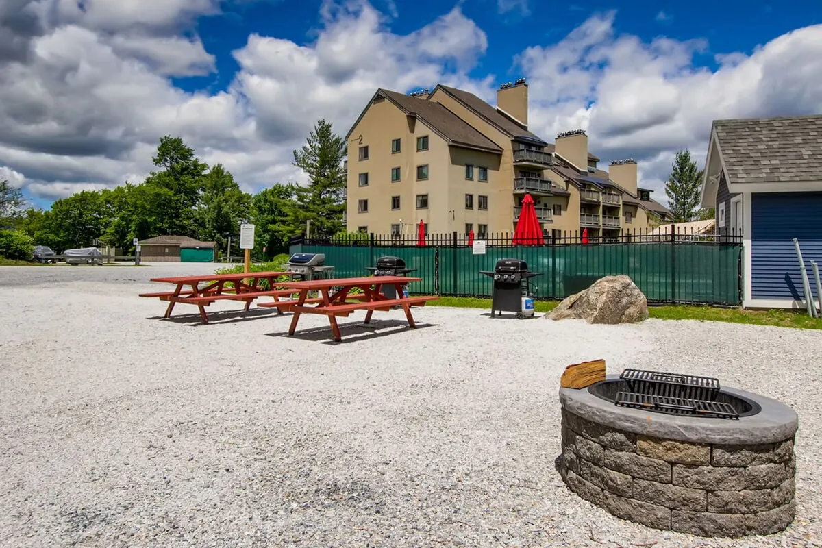 An outdoor space with picnic tables, a fire pit, grill stations, and a building in the background, all enclosed by a green fence. The area is ideal for communal gatherings, barbecues, and fireside evenings during warmer months.
