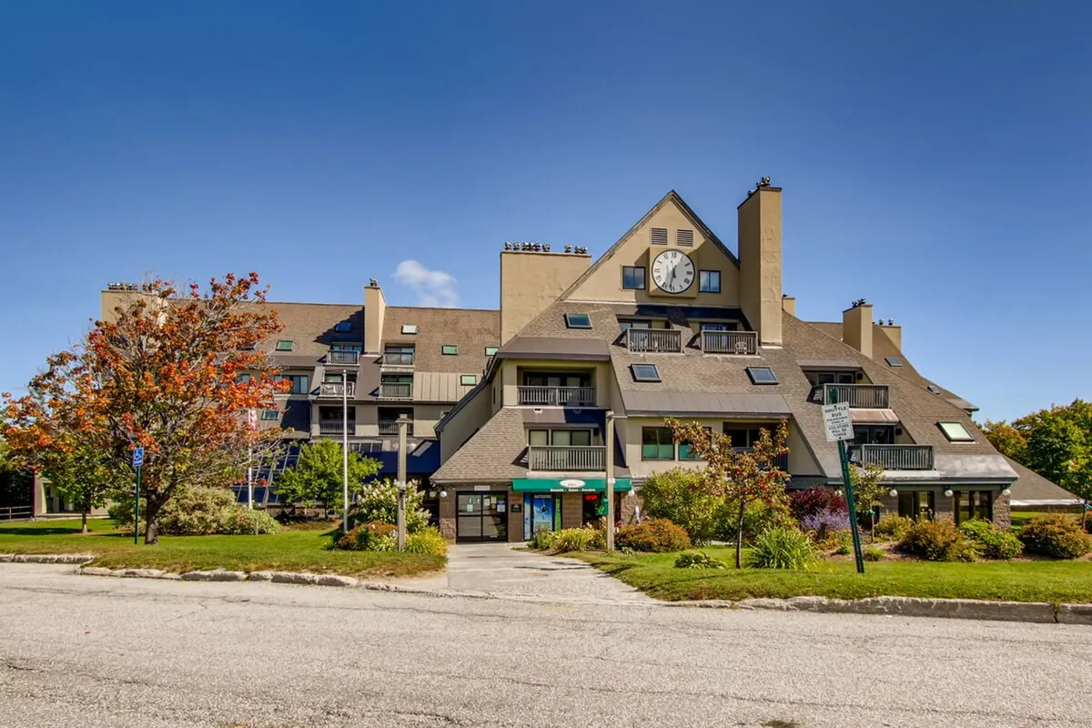An exterior shot highlighting the building’s distinctive architecture, a prominent clock tower, colorful landscaping, and cheerful outdoor gathering spaces. The scene conveys a sense of community and lively resort life.