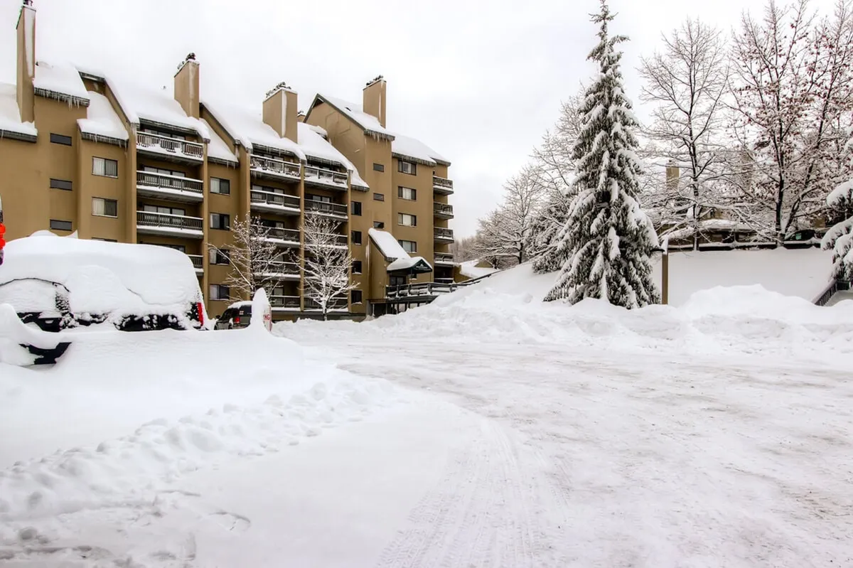 This exterior image captures a snow-covered scene with vehicles parked in front of a multi-story building, framed by tall trees dusted in fresh snow. The tranquil landscape evokes the quintessential winter resort atmosphere, immediately connecting viewers to the experience of waking up right at the heart of Killington's ski area.