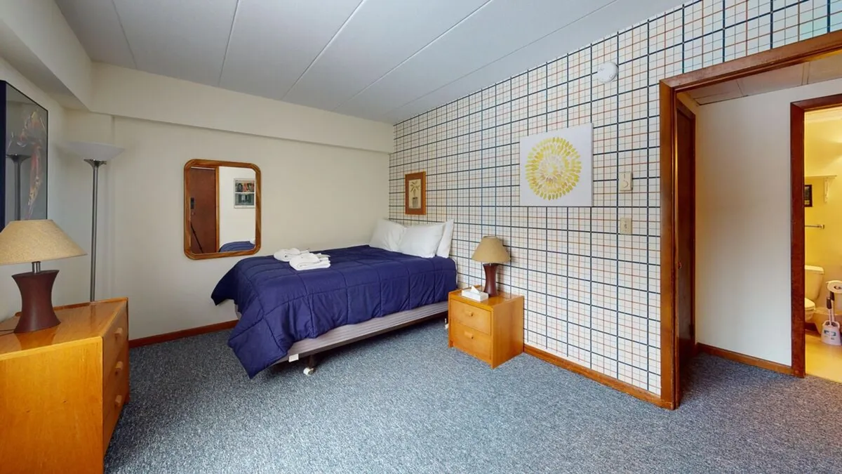 This image displays a bedroom with a plush bed featuring blue bedding, wooden nightstands, a decorative wall art piece above the bed, and a mirror. The soft lighting and cozy arrangement create an inviting atmosphere ideal for restful nights.