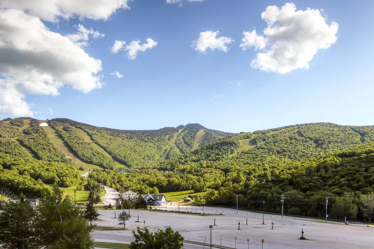 This scenic view showcases the expansive Green Mountain range, rolling green hills, a clear blue sky, and the surrounding landscape near the resort. The image also includes a glimpse of small buildings and an empty parking lot, rooting the adventure in the local setting.