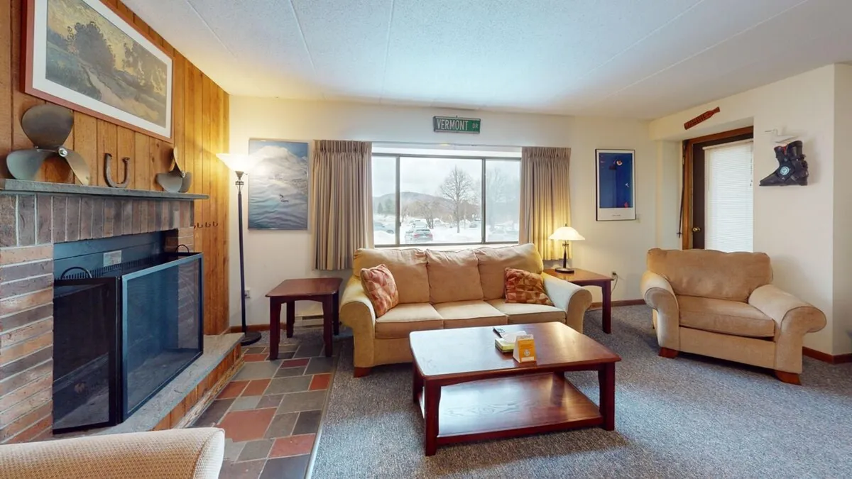 A cozy living area featuring a sofa, a large window that lets in ample natural light, a coffee table, and a rustic fireplace as the centerpiece—even though it's currently inoperable. The setting exudes warmth and offers an inviting space for relaxation, reading, or socializing while watching the snow fall outside.